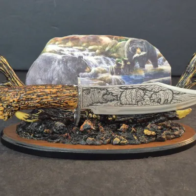 Decorative bowie knife with a faux-antler handle and etched bear scene on the blade, displayed on a matching stand featuring a painted...