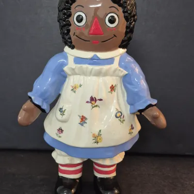 Vintage ceramic African American Raggedy Ann doll figurine with a hand-painted floral apron, blue dress, and striped socks.