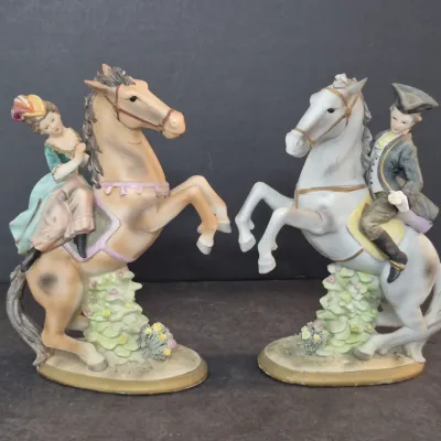 Pair of vintage porcelain figurines depicting a 18th-century style lady and gentleman riding rearing horses, finished in a matte glaze.