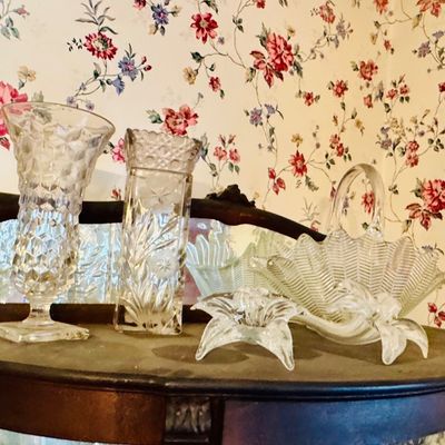 Sale Photo Thumbnail #24: Assorted vintage clear pressed and cut glass tableware, including vases, a basket-style dish, and a small flower-shaped bowl, displayed on a wooden cabinet.