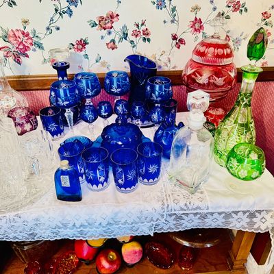 Sale Photo Thumbnail #38: Collection of vintage cut-to-clear glassware, featuring cobalt blue, ruby red, and emerald green decanters, goblets, and tumblers displayed on a lace-covered table.