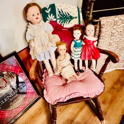 Sale Photo Thumbnail #17: Collection of mid-century vintage dolls, including a larger composition doll and three smaller hard plastic dolls, displayed on a wooden rocking chair with a pink cushion.