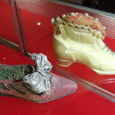 Sale Photo Thumbnail #30: Two vintage novelty shoe pin cushions, including a yellow high-top boot style with gold trim and faux pearls, and a red low-heeled shoe style with a silver bow and green moss-like filling.