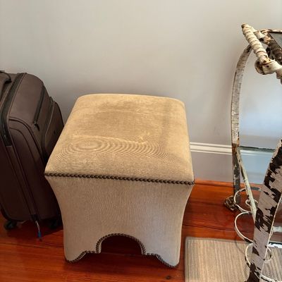 Contemporary beige upholstered ottoman or footstool featuring nailhead trim detailing around the lower edge and seat.
