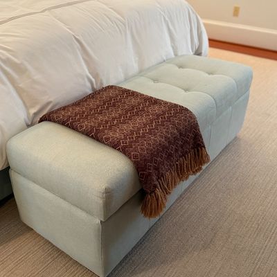 Contemporary tufted light blue upholstered storage ottoman bench draped with a patterned fringe throw blanket.