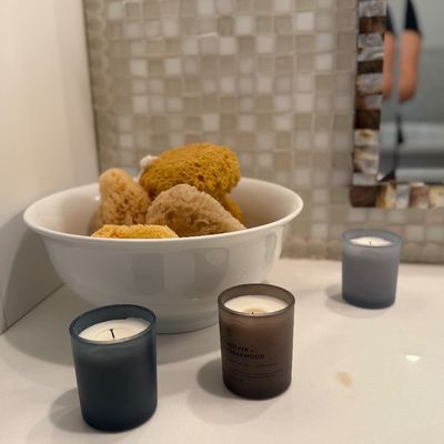 A white ceramic bowl filled with natural sea sponges, displayed alongside three frosted glass votive candles on a bathroom vanity.