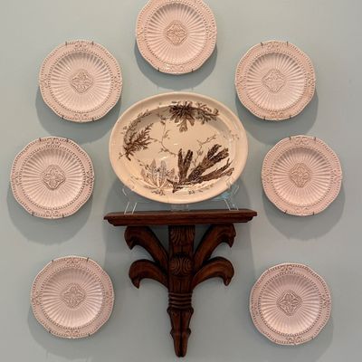 Wall-mounted display featuring seven embossed cream ceramic plates arranged around an antique transferware platter on a carved wooden...