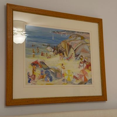 Framed impressionist-style art print depicting a crowded beach scene with colorful umbrellas and sailboats in a warm wooden frame.