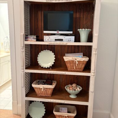 Tall distressed white bookcase featuring shuttered sides, beadboard backing, and decorative trim, displayed with small electronics,...