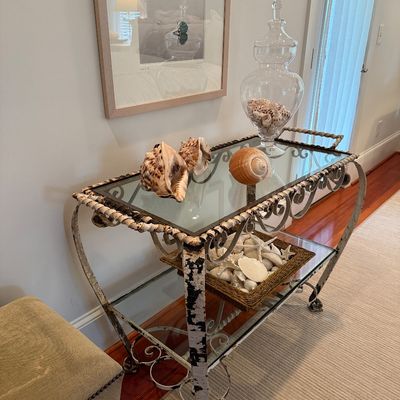 Distressed wrought iron two-tier bar cart with glass shelves, styled with coastal decor including large seashell specimens and a lidded...