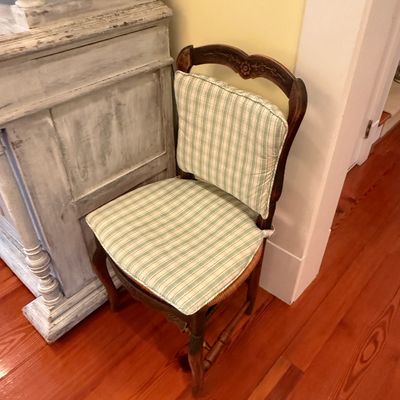 Vintage carved wooden side chair with green and white plaid cushions, positioned beside a distressed whitewashed hutch.
