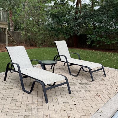 Pair of modern outdoor lounge chairs with dark metal frames and light woven sling fabric, accompanied by a small round glass-top side table.