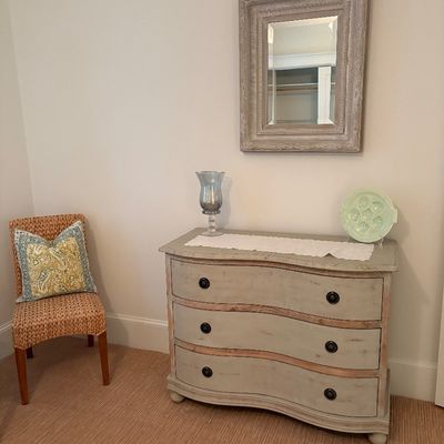 French Country style distressed gray serpentine-front three-drawer dresser, paired with a matching framed wall mirror and a woven accent...