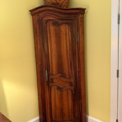 French Provincial style corner cabinet in a warm wood finish, featuring ornate carved accents on the single door and topped with a woven...