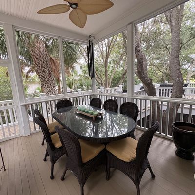 Outdoor wicker patio dining set featuring an oval glass-top table surrounded by eight dark brown wicker armchairs with tan seat cushions...
