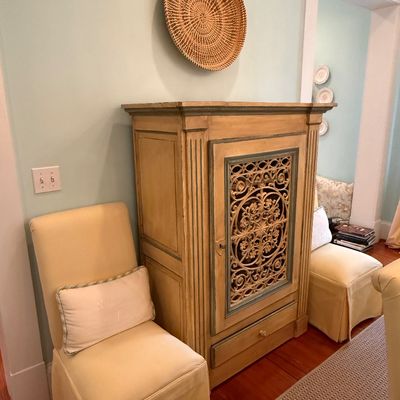 Sale Photo Thumbnail #105: Tall painted wooden armoire with a decorative carved scrollwork door insert, flanked by a pair of cream-upholstered side chairs with skirted bases.