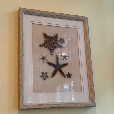 Framed coastal wall art featuring embroidered or beaded starfish designs on a neutral fabric background, presented in a light wood-tone...