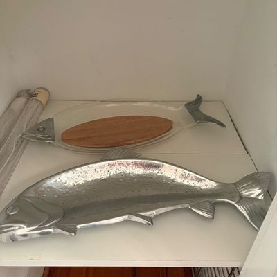Two fish-shaped serving trays on a white shelf, including a large cast aluminum platter and a glass dish with metal head and tail accents.
