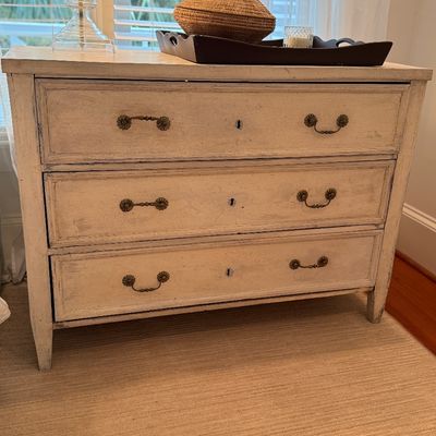 Distressed cream-painted three-drawer chest featuring brass bail handles and keyholes, styled in a French provincial or Swedish gustavian...