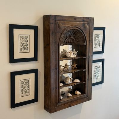 Wall-mounted rustic wooden display cabinet with carved arched top and mirrored back, filled with seashells and flanked by four framed...