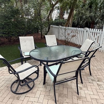 Sale Photo Thumbnail #108: Outdoor patio dining set featuring an oval glass-top table with dark metal frame and six matching sling-back chairs, arranged on a brick paver patio.