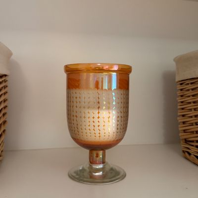 Amber iridescent goblet-style glass hurricane candle holder featuring a geometric patterned center band, displayed on a shelf.