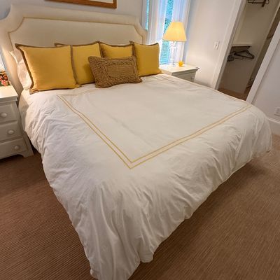 Upholstered king-size bed with a off-white fabric headboard, dressed in white bedding with yellow accent pillows and a matching throw...