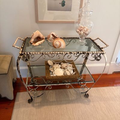 Distressed wrought iron two-tier rolling tea cart with glass shelves, styled with decorative seashells and a glass apothecary jar.