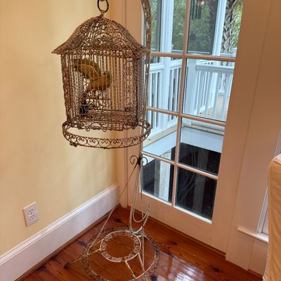 Vintage wire birdcage on a matching wrought-iron floor stand, featuring intricate scrollwork detail and a decorative bird inside.