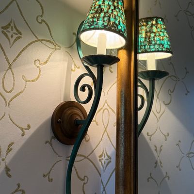 Wall-mounted electric candle sconce with a scrollwork arm and a green mosaic glass shade, reflected in an adjacent mirror.