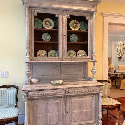 French Country style whitewashed wooden hutch featuring glass-front upper cabinets, carved details, and lower storage, filled with...