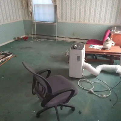 Portable air conditioner with exhaust hose, shown alongside an office chair, a wooden coffee table, and other household items in a room...