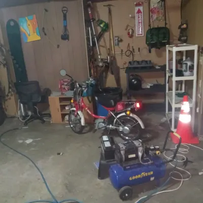 Vintage red moped parked in a garage alongside a Goodyear air compressor, shelving units, and various tools.