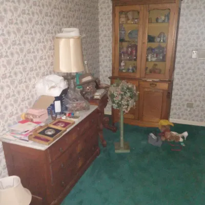 Vintage wooden hutch with glass display doors alongside a marble-top three-drawer dresser, set against floral wallpaper and green carpeting.