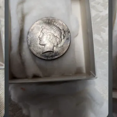 Screenshot of a mobile phone displaying a photo of a 1923 Peace silver dollar coin resting on cotton padding inside a small box.