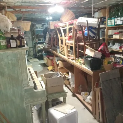 Cluttered basement storage area featuring a green-painted upright piano, wooden dining chairs, shelving units, and various household items.
