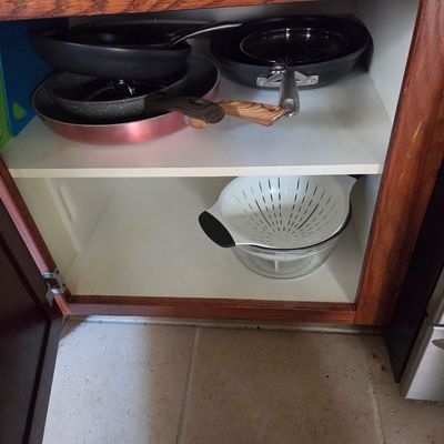 Sale Photo Thumbnail #77: Assorted non-stick frying pans and a white plastic colander stored inside a wooden kitchen cabinet.