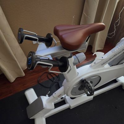 Sale Photo Thumbnail #8: NordicTrack stationary exercise bike in white, featuring a brown leather-style seat and integrated dumbbell holders with weights.
