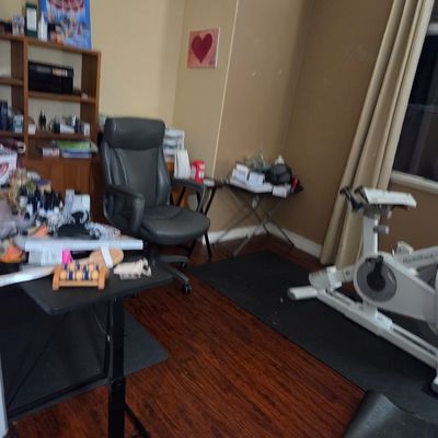 Sale Photo Thumbnail #12: Home office and exercise space featuring a grey leather rolling office chair, a white stationary exercise bike, and various small tables and shelving units.