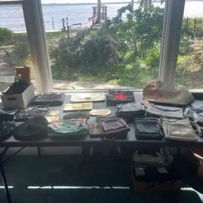 Collection of vintage and modern women's handbags, clutches, crossbodies, and hats displayed on a folding table in front of a window...