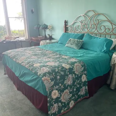 King-size bed featuring an ornate scrollwork metal headboard, dressed in teal bedding with a floral quilt runner and a burgundy bed skirt.