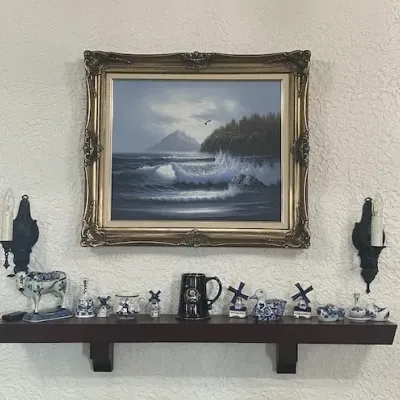 Framed oil painting of a stormy seascape hanging above a mantel lined with blue and white porcelain decor and a dark glazed mug.