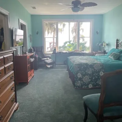 Spacious bedroom featuring wooden dressers, a bed with teal bedding, an upholstered armchair with ottoman, and large windows with a...