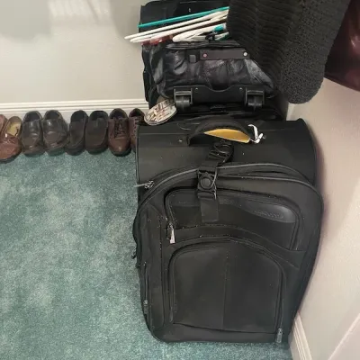 Collection of black rolling luggage bags stacked in a closet, with plastic clothes hangers on top and men's dress shoes lined up...