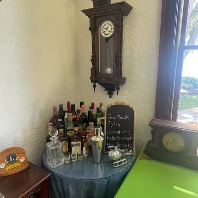 Antique carved wood wall clock mounted above a glass-top bar table setup with assorted liquor bottles, a crystal decanter, and a...