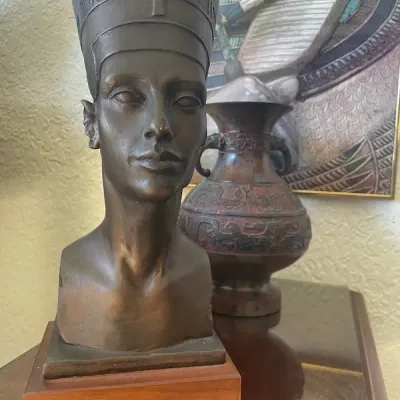 Egyptian-revival bronze bust of Nefertiti mounted on a wooden base, displayed alongside a metal vase with embossed patterns.