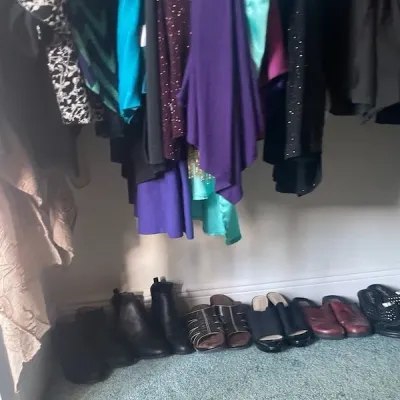 Selection of women's shoes and hanging clothing inside a closet, including ankle boots, mules, slides, and various dresses and tops.