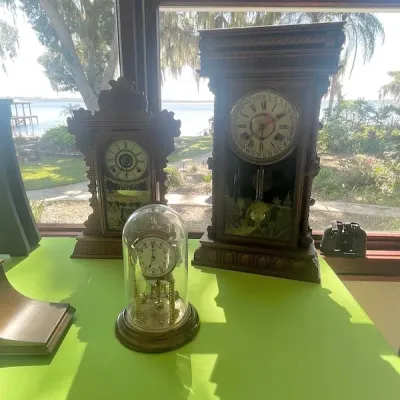 Collection of three antique mantle clocks, including a brass anniversary clock under a glass dome and two wooden pendulum shelf clocks on...