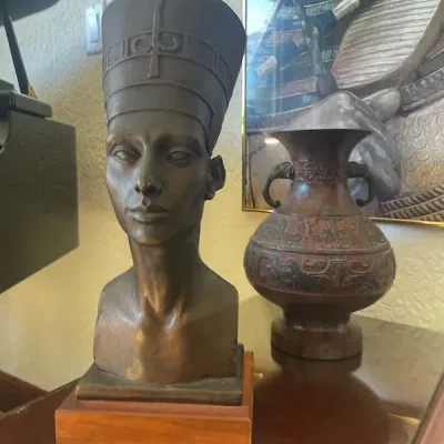 Bronze bust of Egyptian Queen Nefertiti mounted on a wooden base, displayed alongside  a metal vase with embossed patterns.