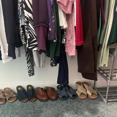 Assorted women's clothing hanging on a rack above a row of various sandals, clogs, and slip-on shoes.